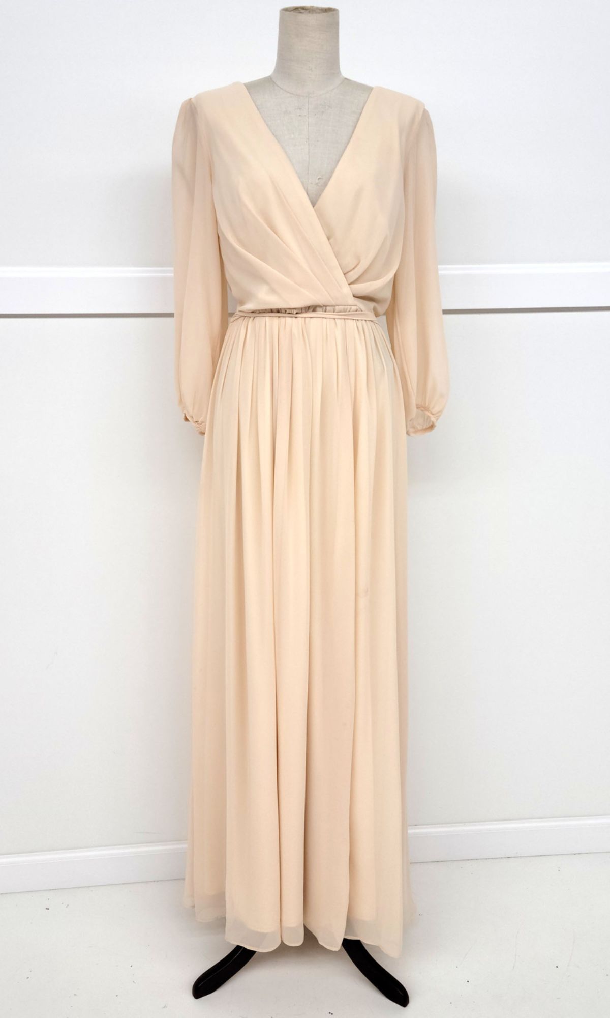 The Chicas J012B, a long-sleeve modest formal dress in light beige chiffon with a deep V-neck and gathered waist, is displayed on a dress form against a white background.